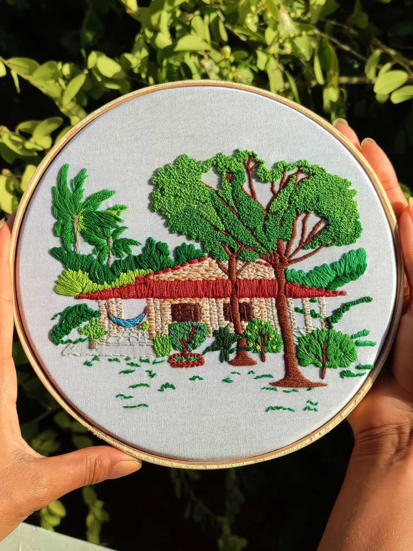 Bastidor com casa de telhado vermelho, rede e árvore bordadas em verde, erguido entre as folhas do quintal
